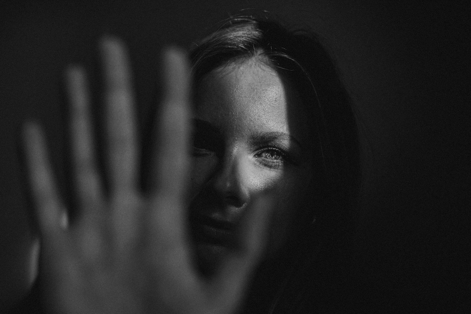 Black and white portrait of a woman with her hand raised in front of her face, one eye visible through the gap — representing concealment and the choice to be seen